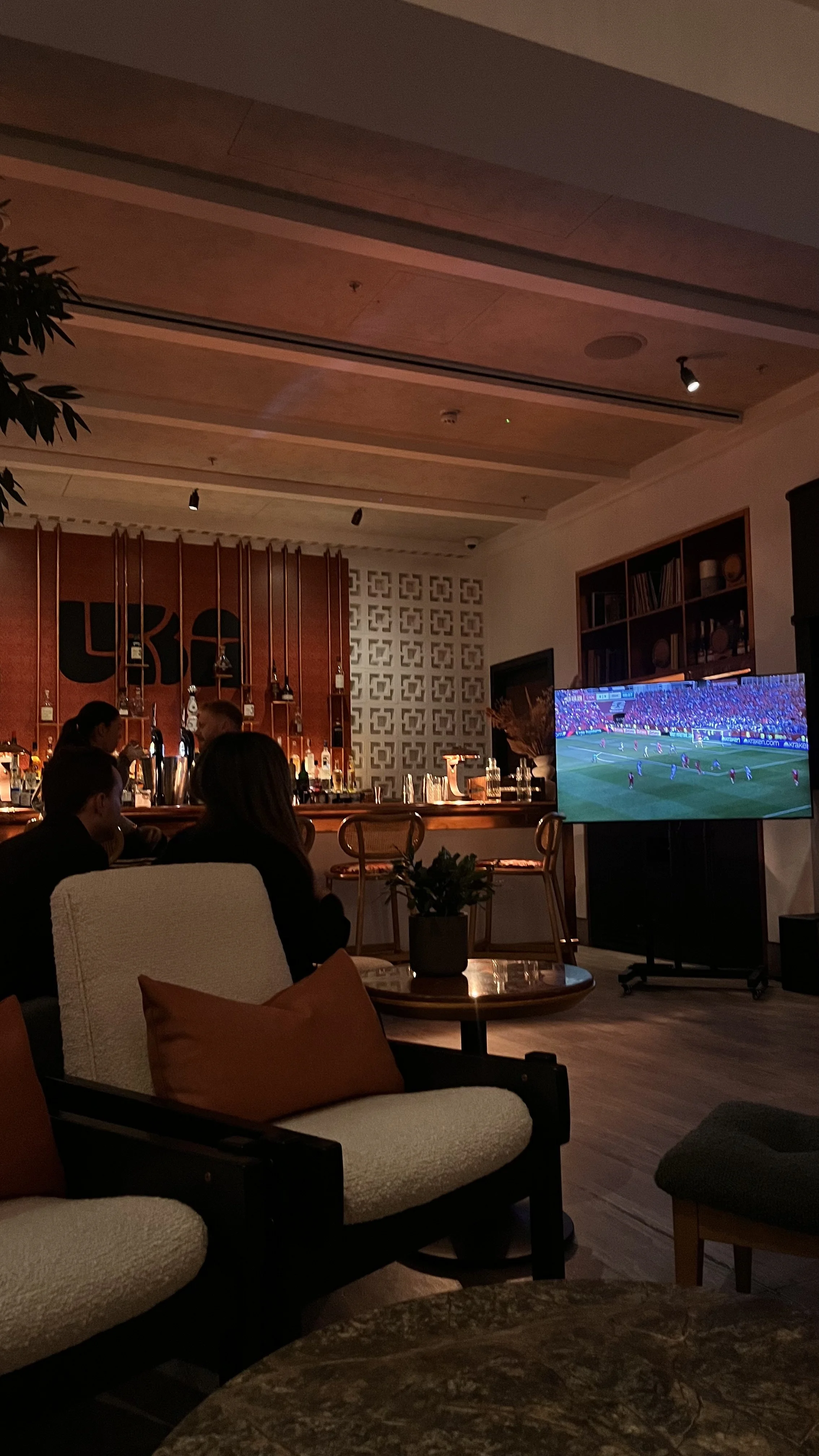 A dimly lit bar area with people watching a soccer game on a TV. The bar has various bottles and glasses, and the seating includes plush chairs and a small round table with a plant.