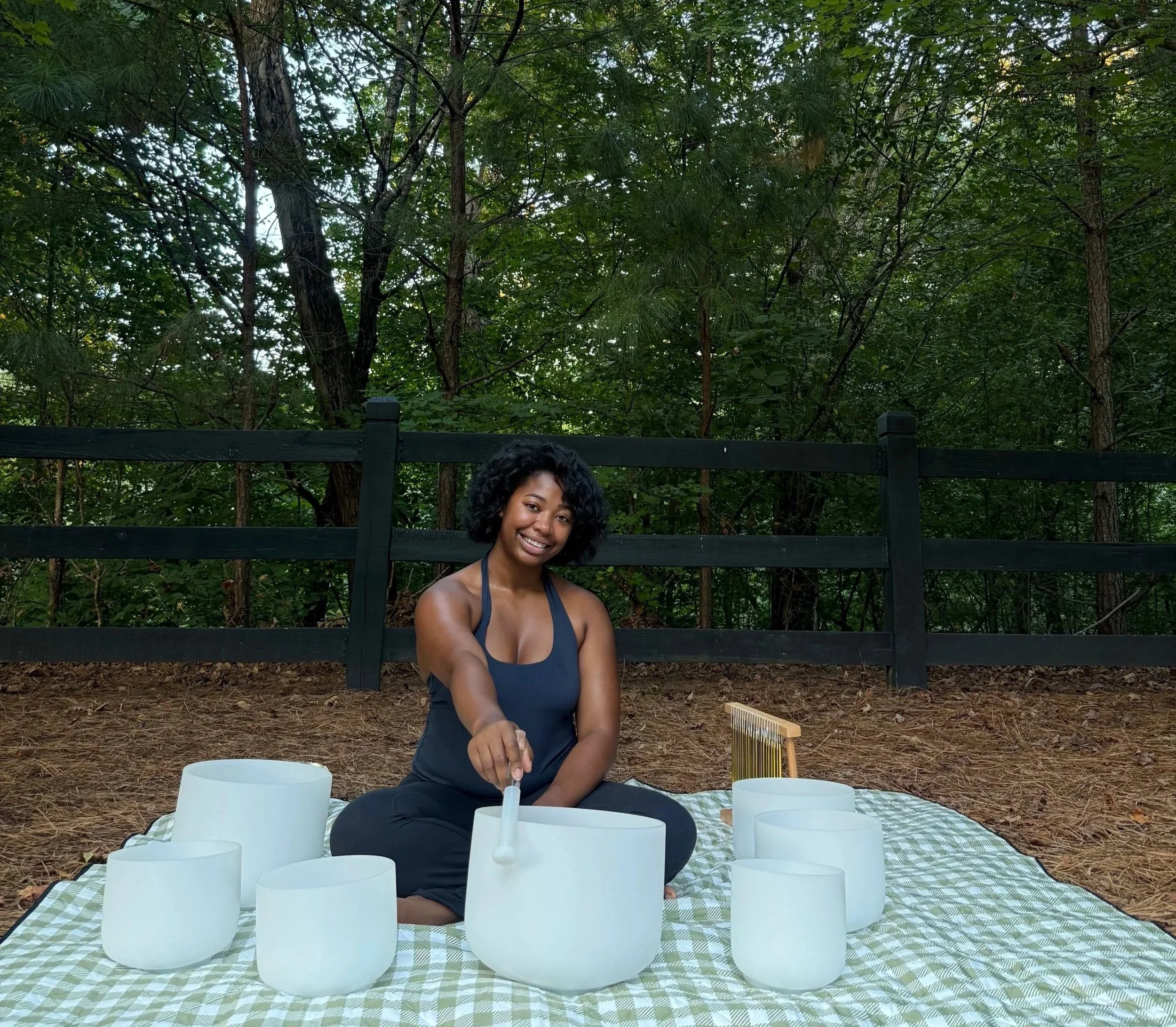 A young woman sitting outdoors at a table with white ceramic singing bowls and a scratching instrument, surrounded by trees and greenery.