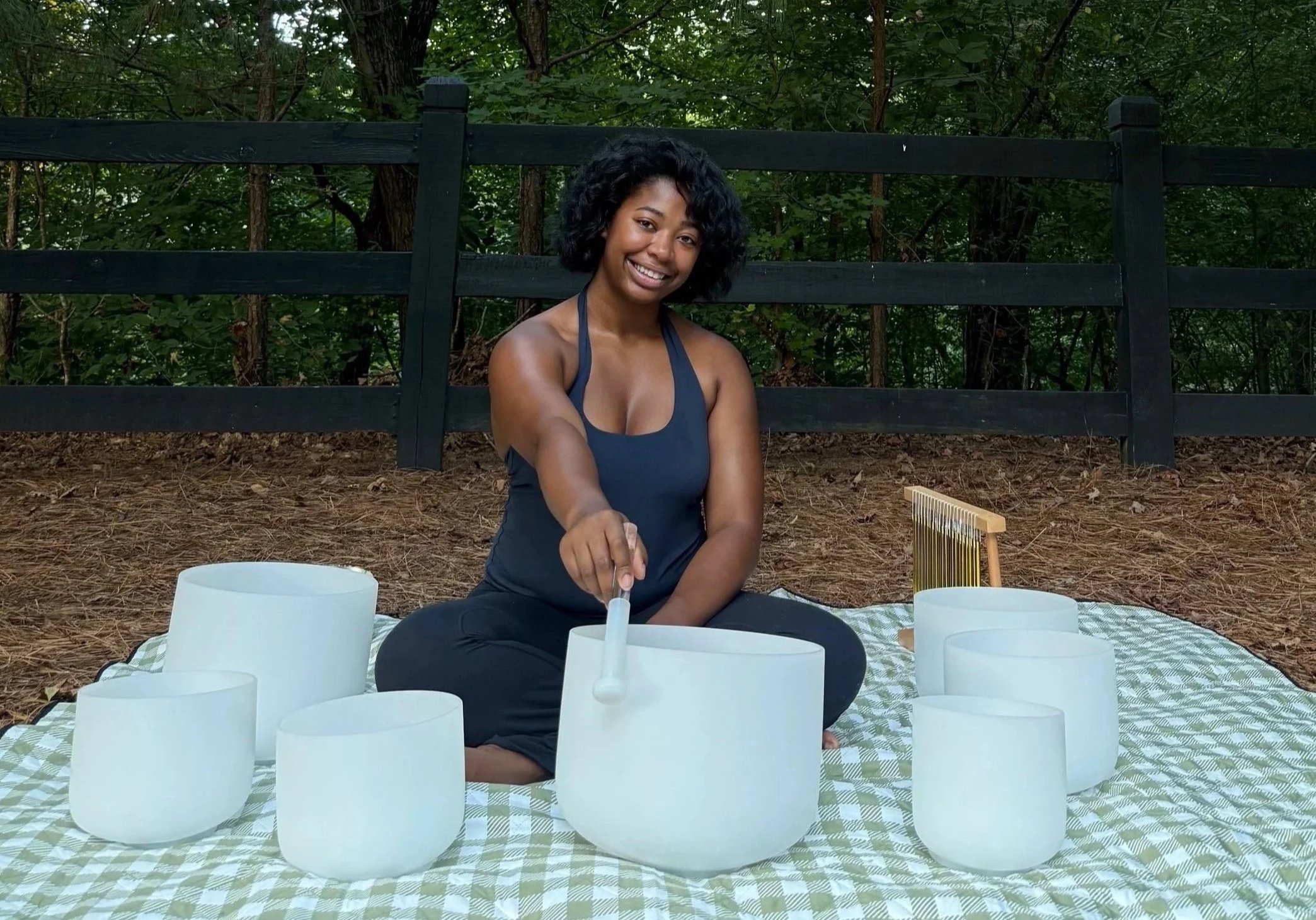 A woman with curly black hair and a dark blue tank top sitting outdoors on a checkered blanket, playing singing bowls surrounded by white bowls and a golden handpan, with a black wooden fence and green trees in the background.