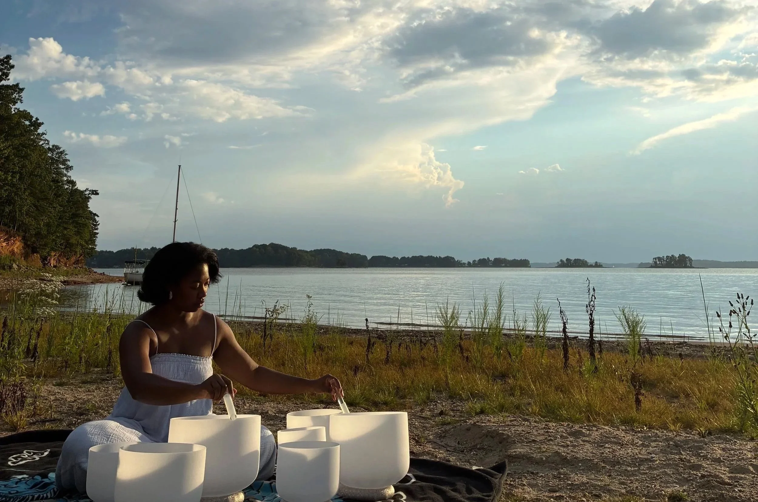 Gracelyn sits on a blanket on a sandy lakeshore in Georgia, playing crystal singing bowls and rain drums as the sun shines through the clouds over the water.