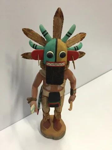 A figurine of a humanoid creature with a colorful, decorated tribal headdress featuring feathers and beads, standing on a wooden base.