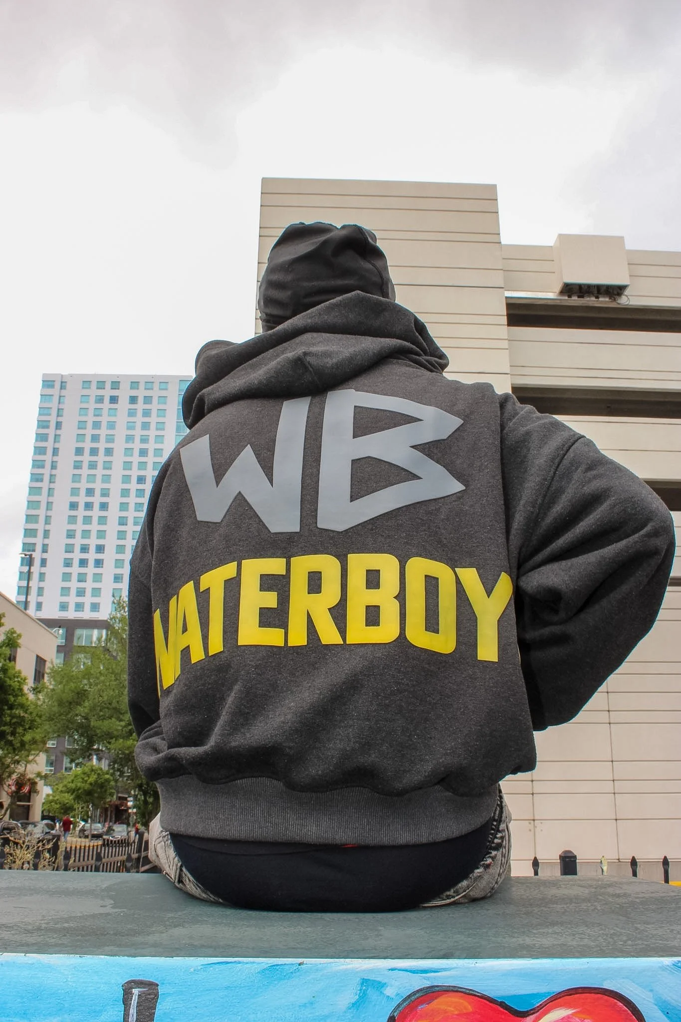 Person sitting outdoors at a cityscape, wearing a black hoodie and a gray sweatshirt with yellow text, with tall buildings and cloudy sky in the background.
