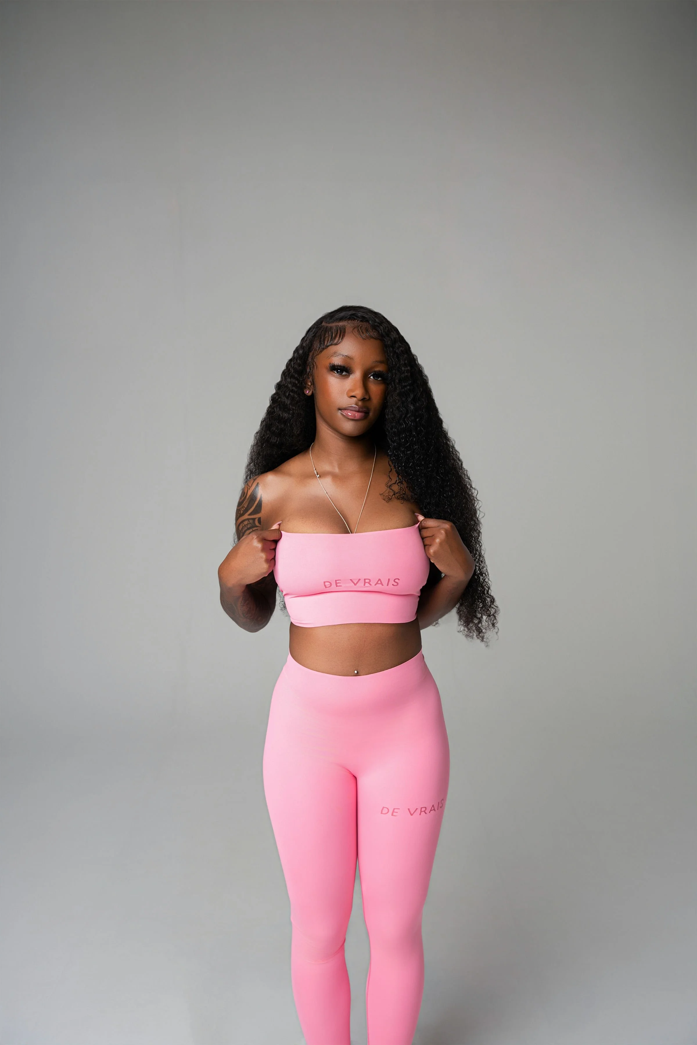 A woman with long curly black hair, wearing pink activewear with the phrase 'DE VRAIS' printed on the top and leggings, poses against a plain gray background.