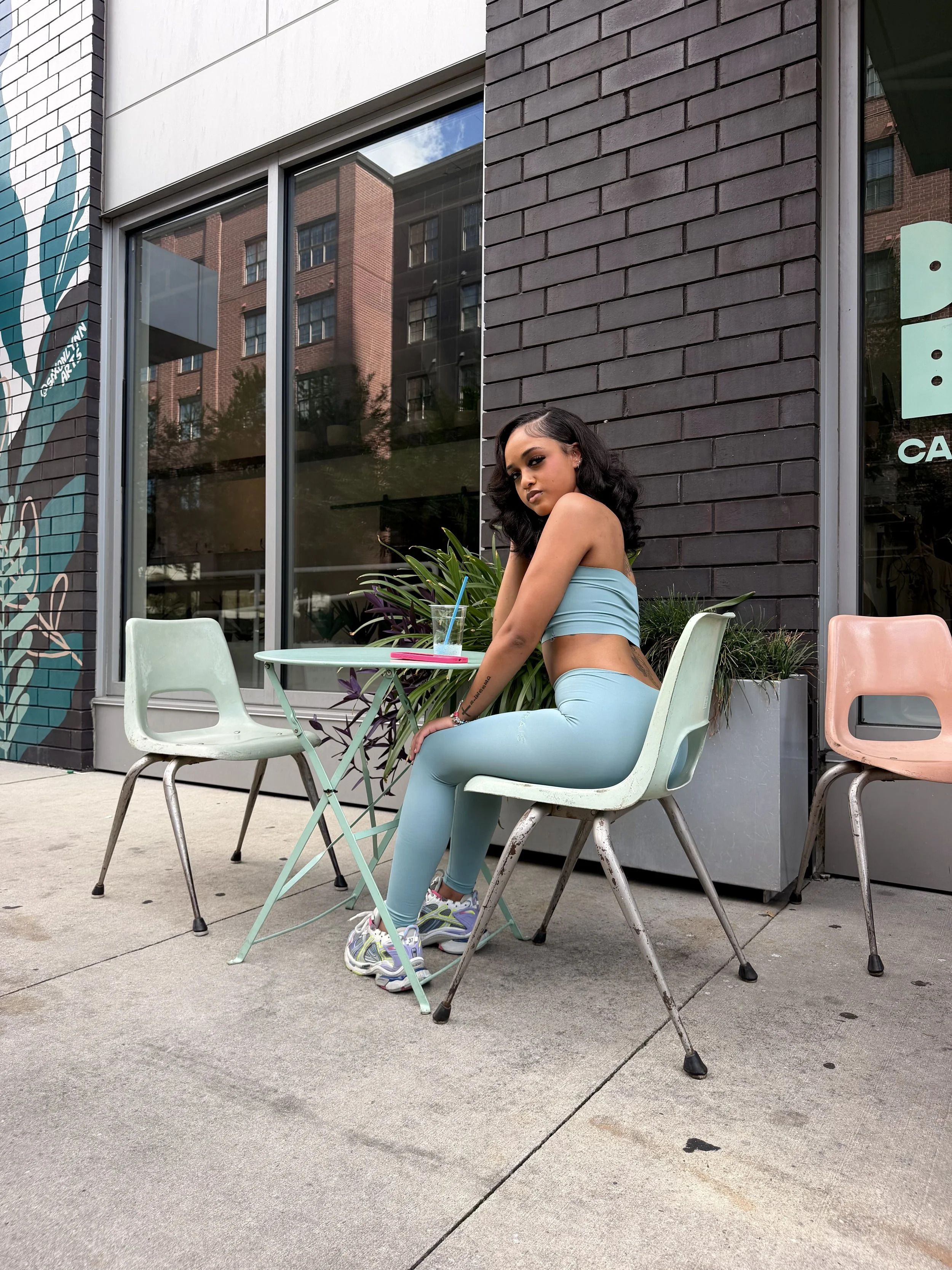 A woman with dark wavy hair, wearing a light blue workout outfit and colorful sneakers, sitting at a small pastel table outside of a brick building with large windows.