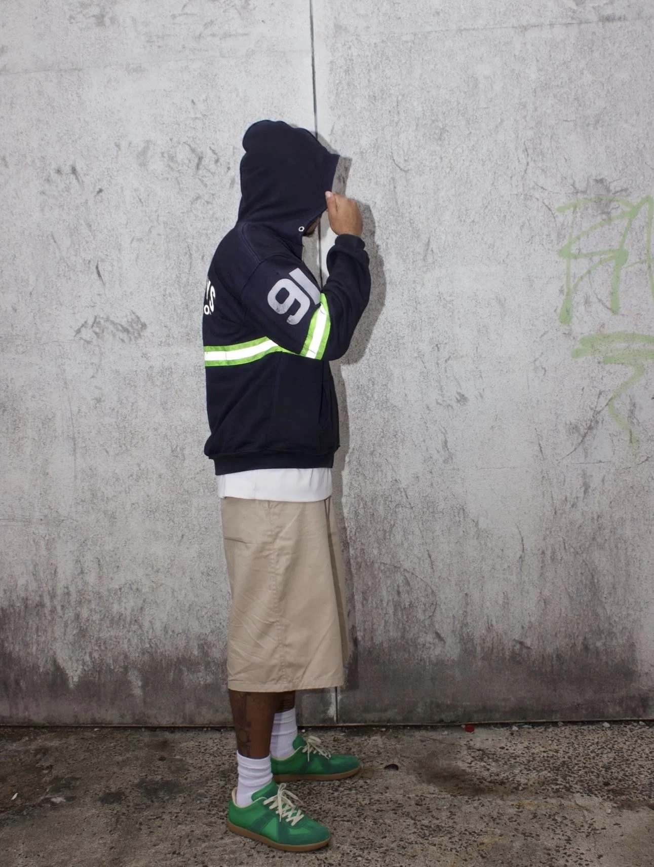 A person standing against a concrete wall, wearing a black hoodie, beige shorts, green sneakers, and white socks, with the hoodie pulled over their head and their face obscured.