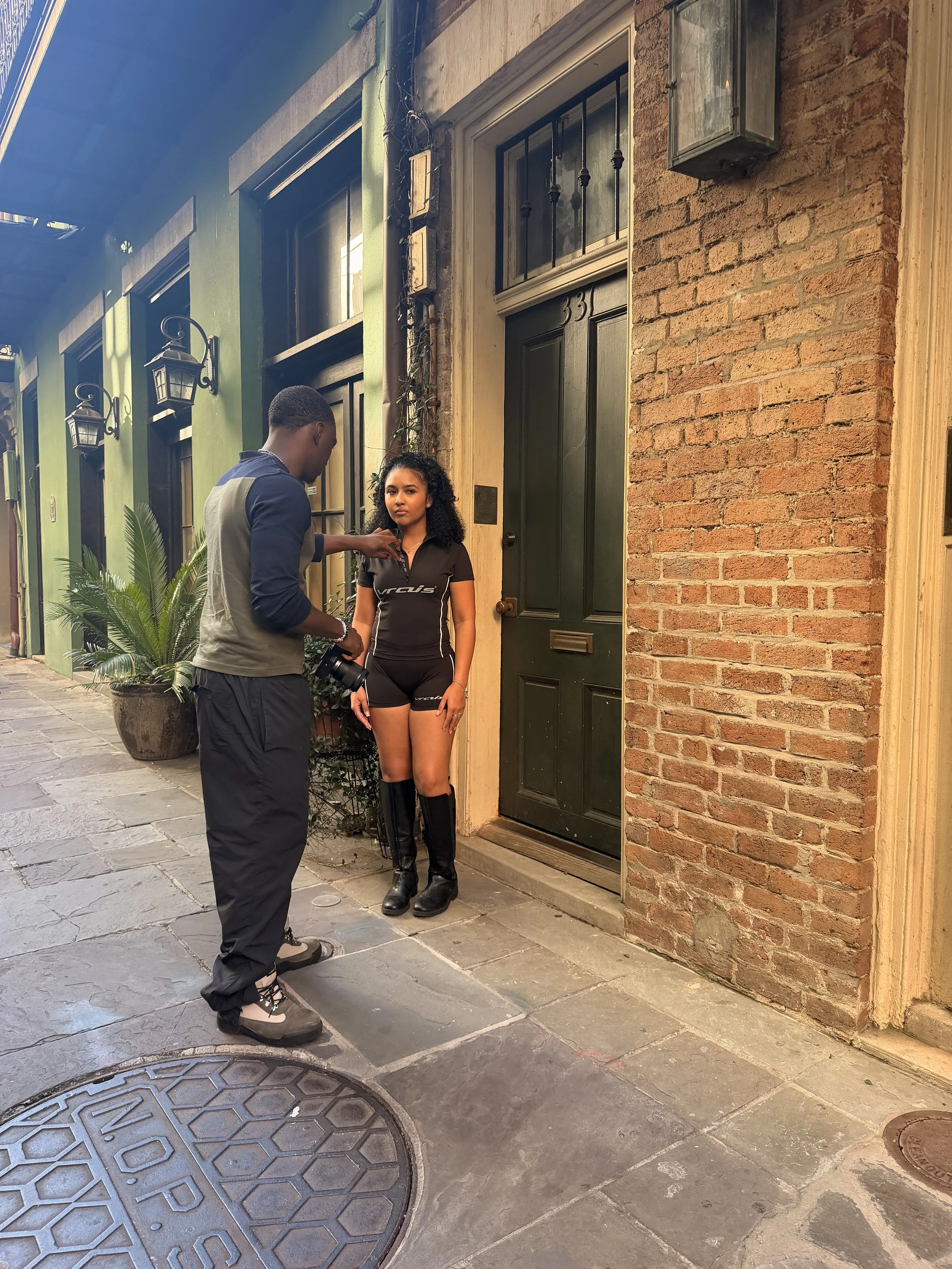 A man and a woman standing outside a brick building with a green door and window. The man is taking a photo or video of the woman, who is looking at the camera with a neutral expression. The woman is wearing a black sports outfit and black boots, and the man is dressed casually with a camera hanging from his neck.