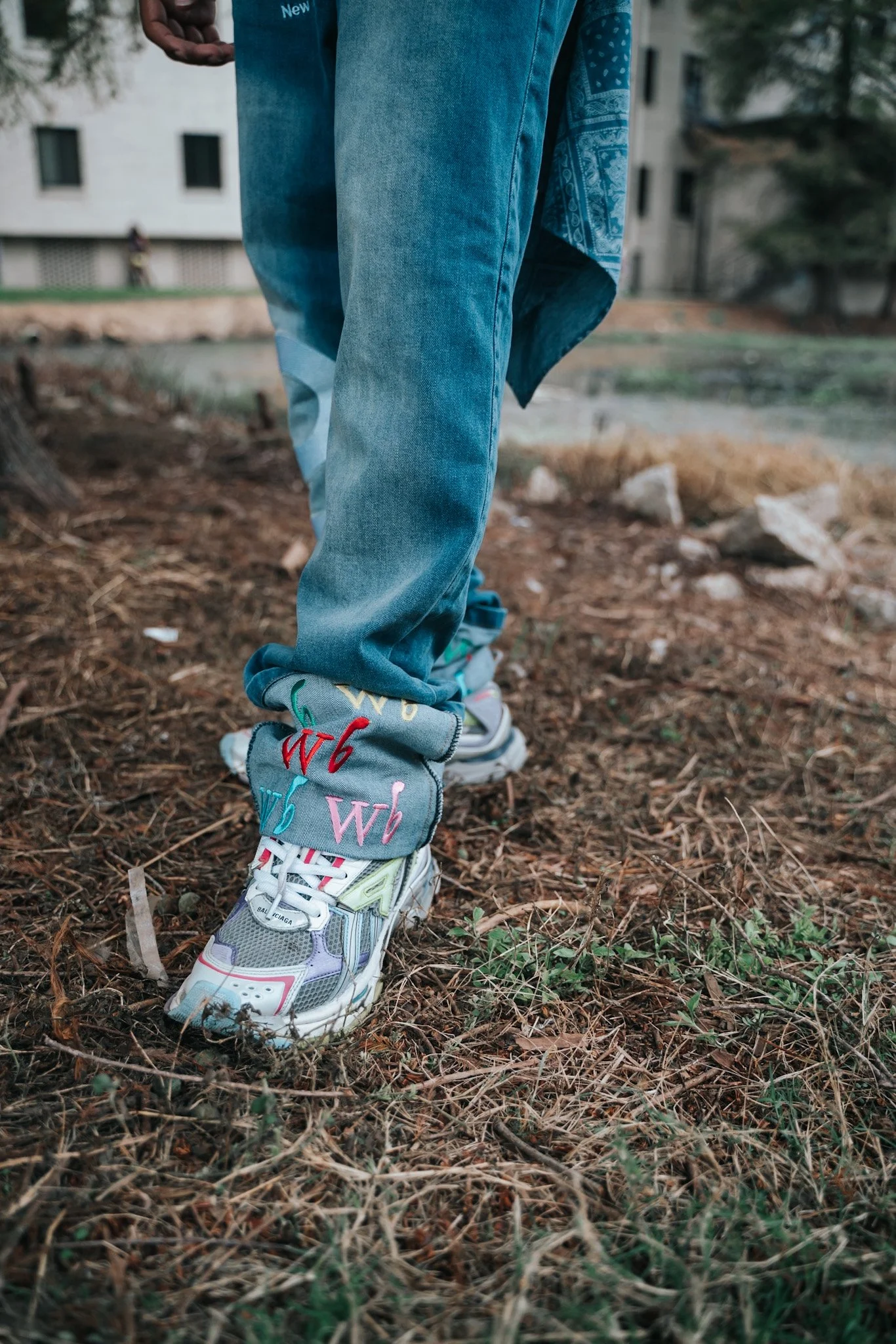 Close-up of a person's legs wearing baggy blue jeans and colorful sneakers, standing on dirt and grass.