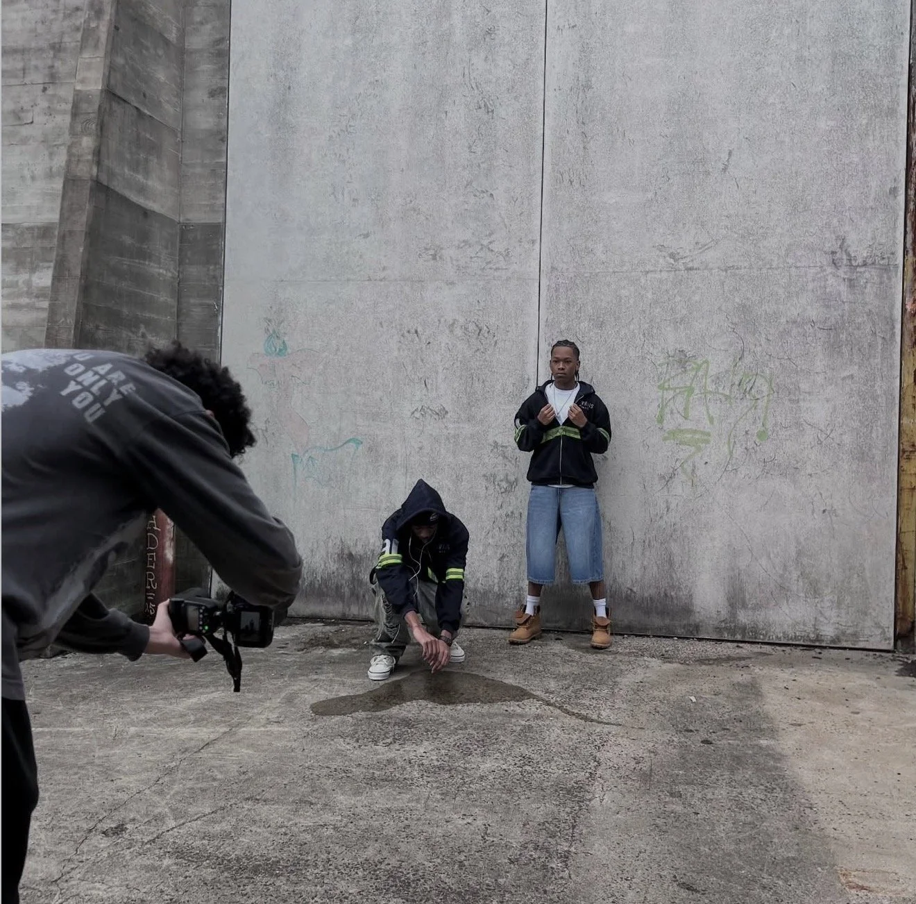 Two young men standing against a large concrete wall, one squatting and touching the ground while the other stands with fists raised, and a person in the foreground taking a photo with a camera.