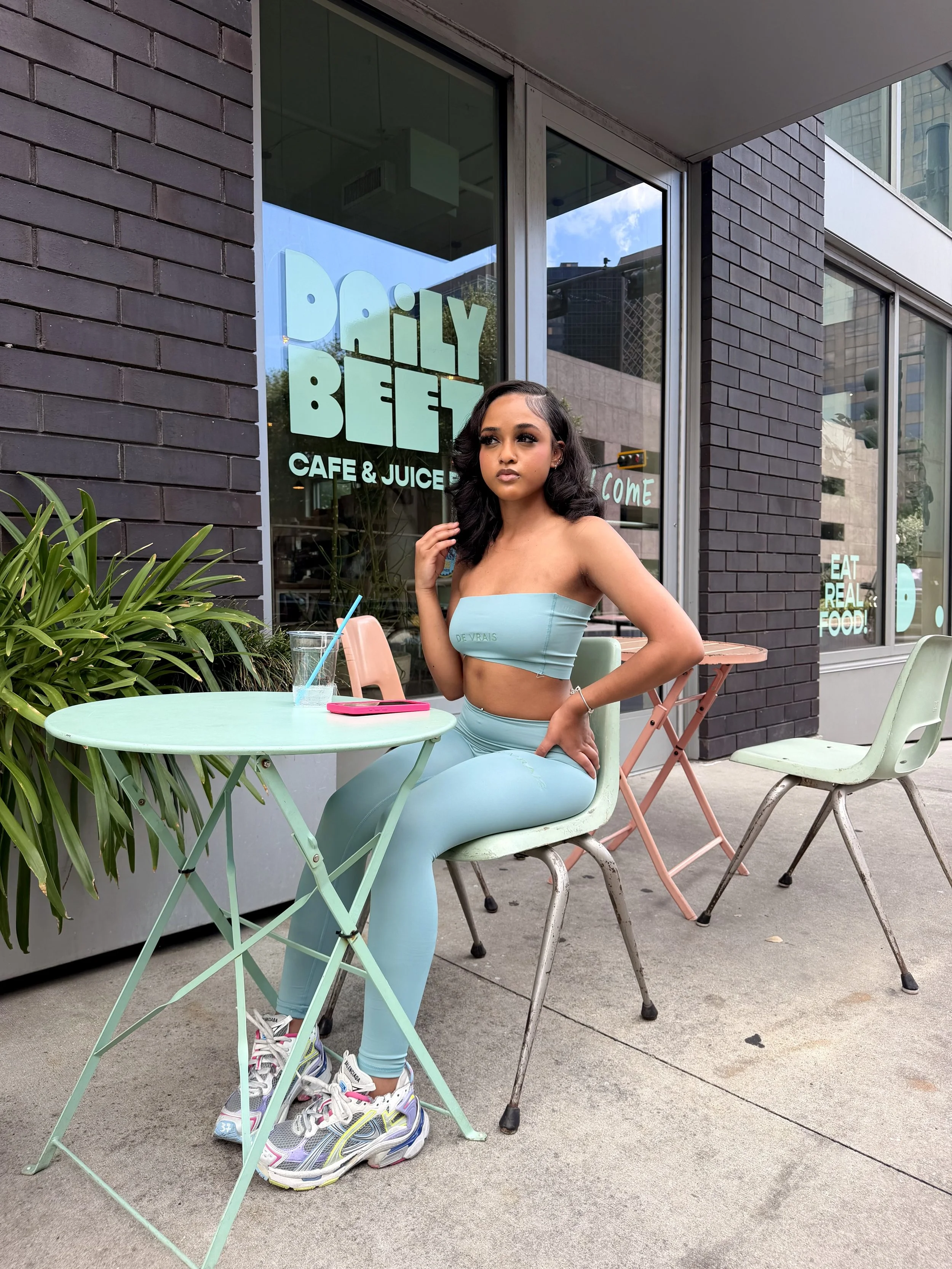 A woman with black wavy hair sitting at an outdoor café table, wearing a light blue strapless top and matching leggings, with sneakers. The table has a pink phone and a glass with a straw. Behind her is a glass window with green and white text for a café called Daily Beetz, and some greenery. The sidewalk has some chairs and plants.