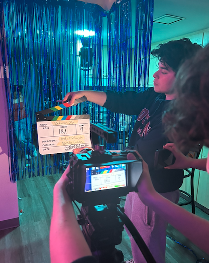 A young woman with short black hair wearing a black hoodie is holding a clapperboard in a room decorated with blue metallic streamers, while someone else is filming her with a professional camera.