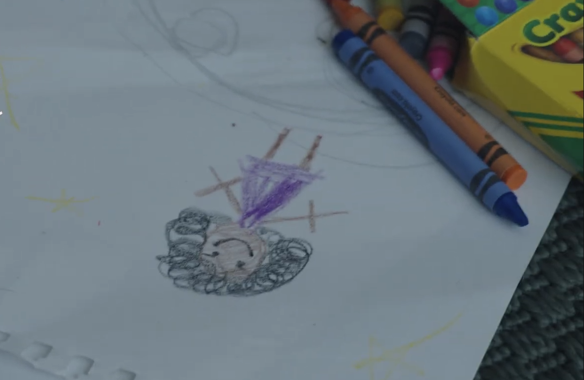 Child's drawing of a person with curly hair, a purple dress, and a happy face. Crayons and a box of Crayola crayons are nearby.