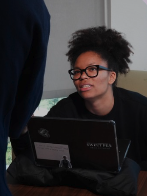 Young woman with glasses and curly hair looking at person with laptop labeled 'Sweet Pea' in an office setting.