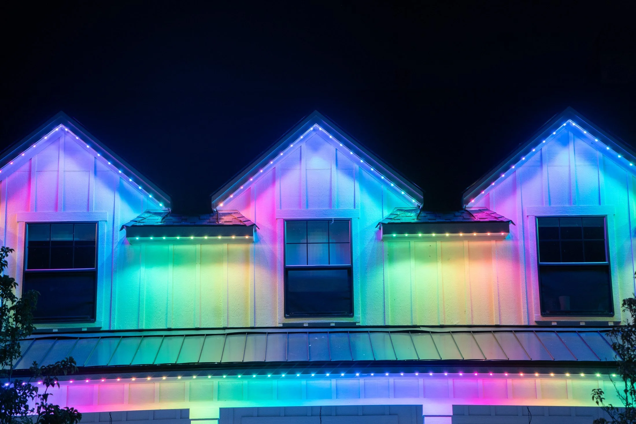 Blue, purple, pink, and green permanent lights illuminate the upper gables and porch eaves of a white-sided home.