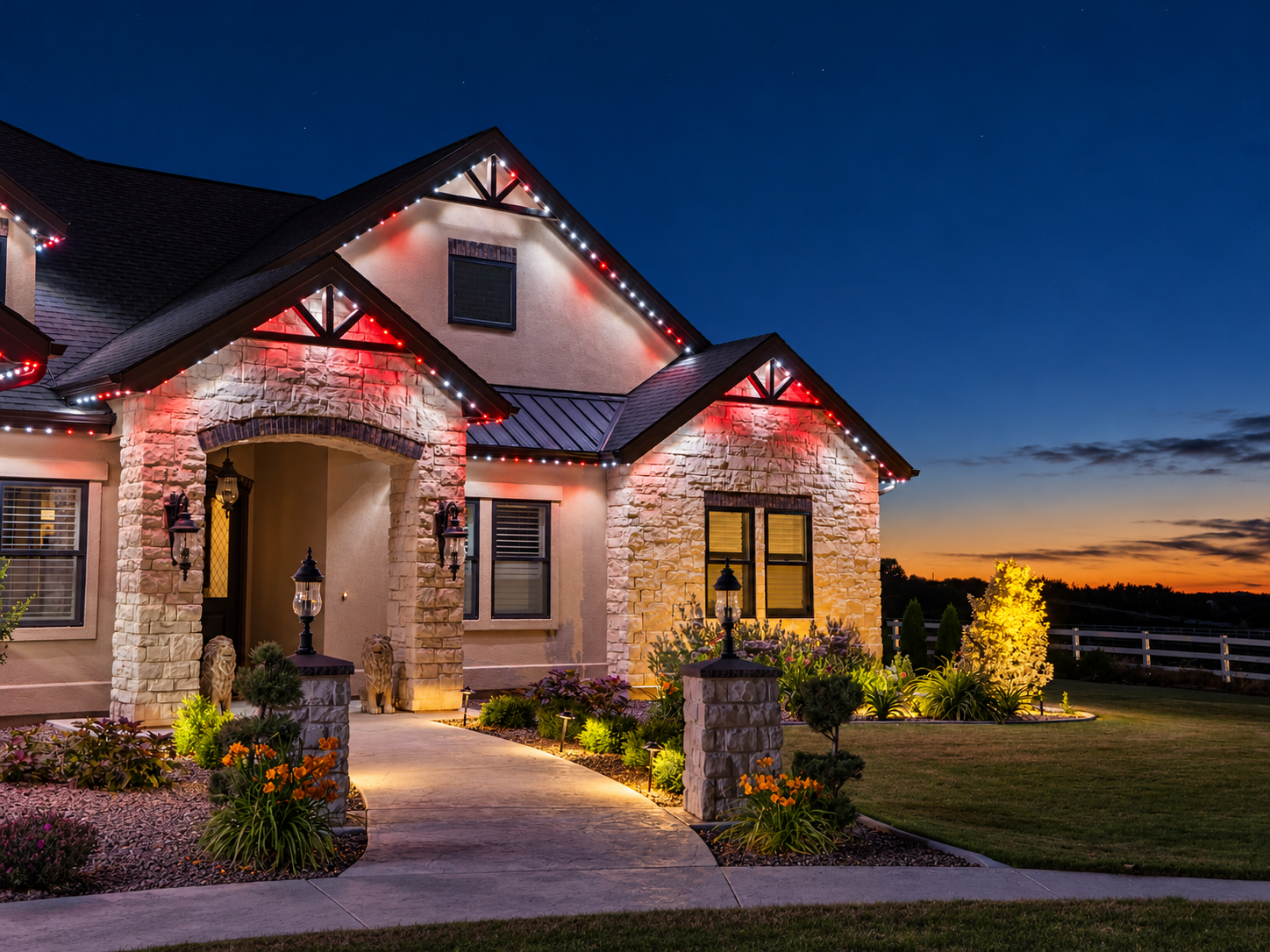 White and red accent lights highlight a stone home’s covered entrance and gables at sunset.