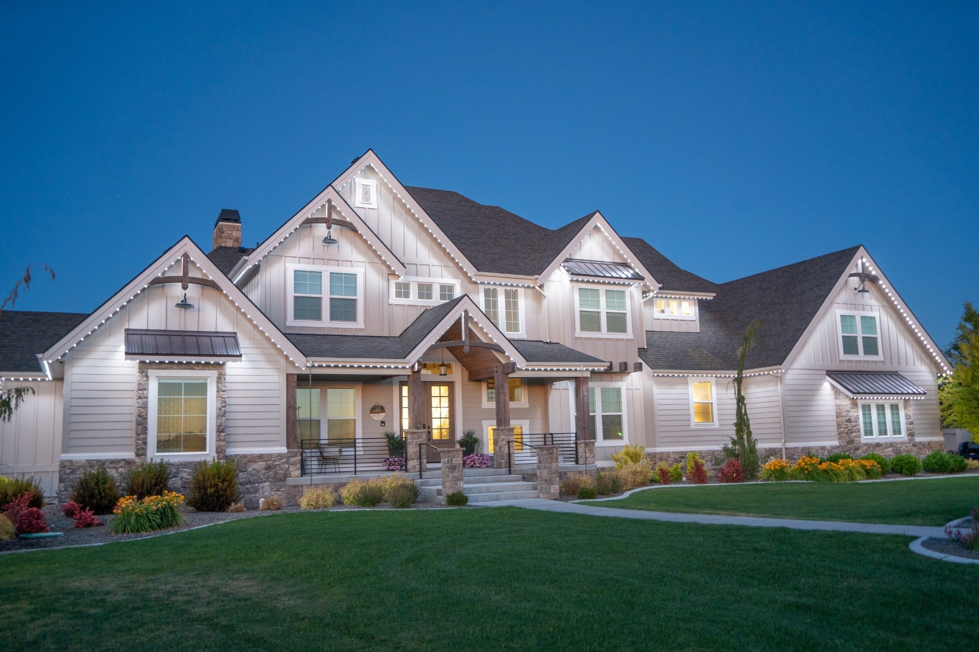 White permanent lighting highlights the upper gables and covered porch of a gray-sided home against an evening sky.