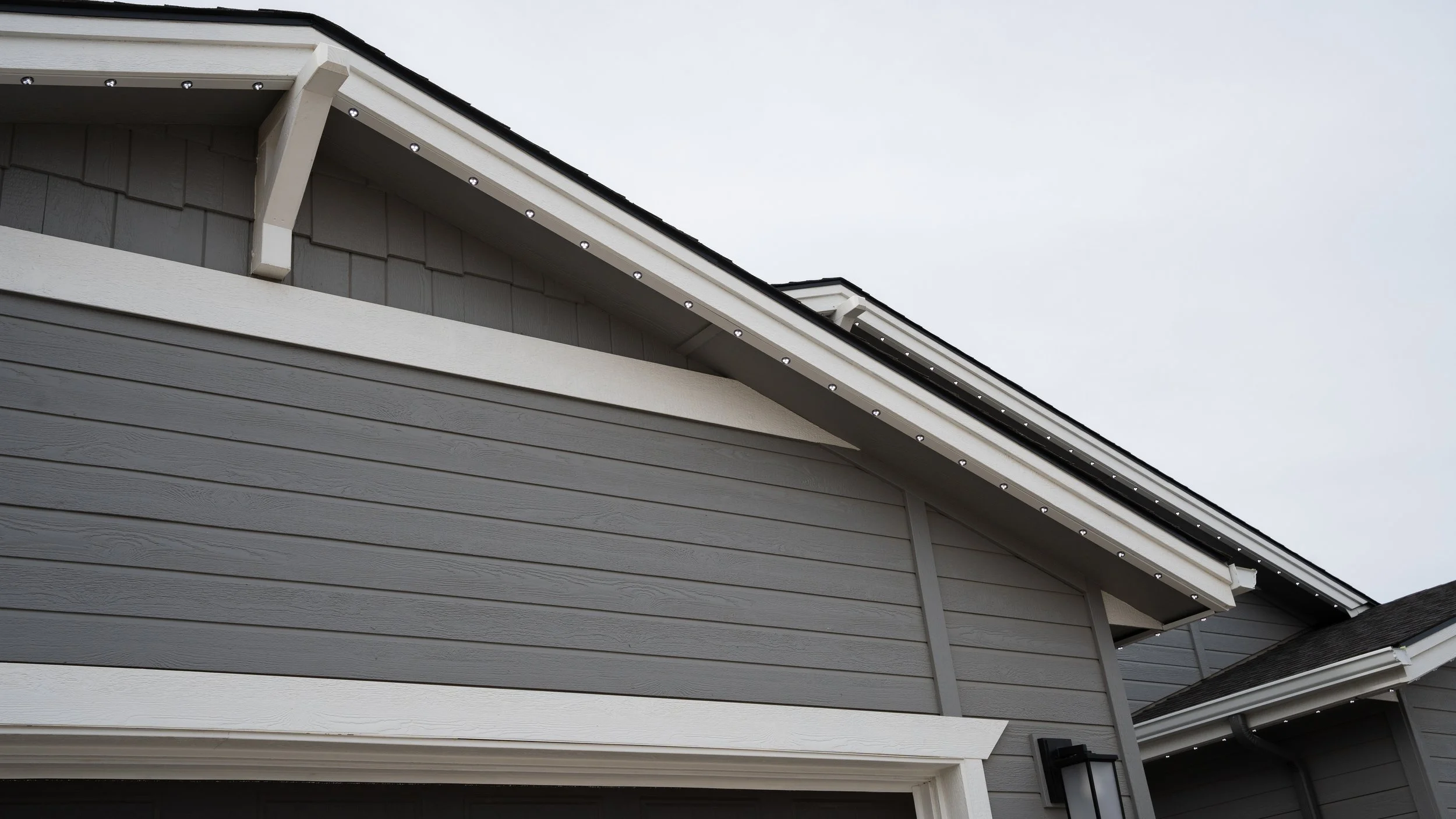 Low-profile permanent lighting beneath the white roof trim of a gray home, shown during daylight.