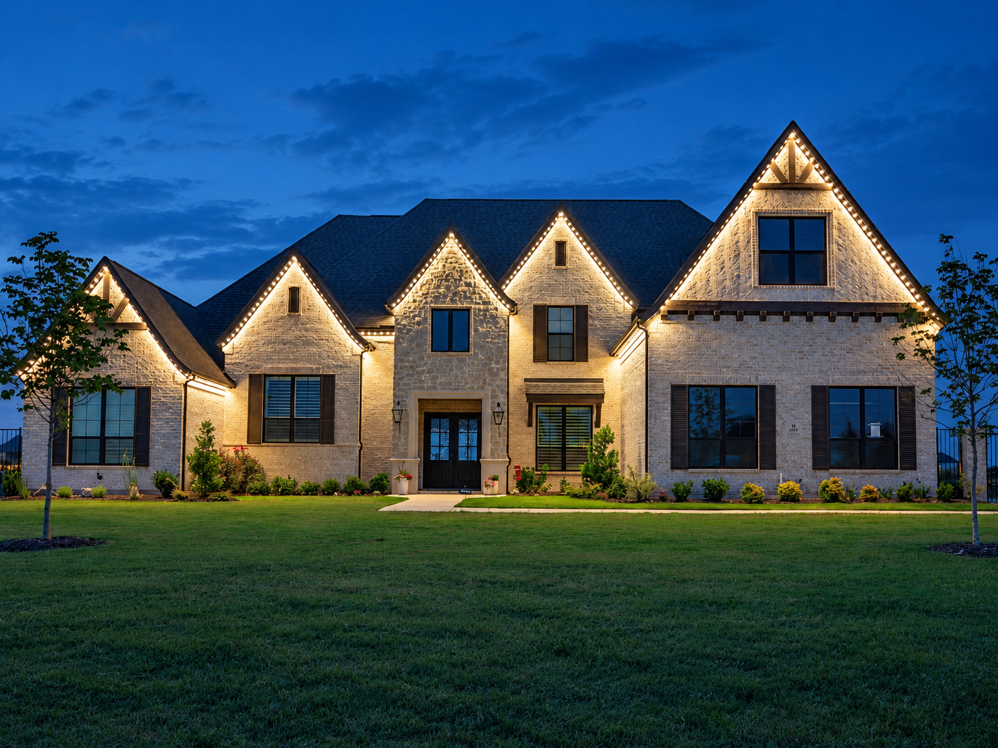 Warm white permanent lighting outlines the peaked gables of a stone home at dusk.