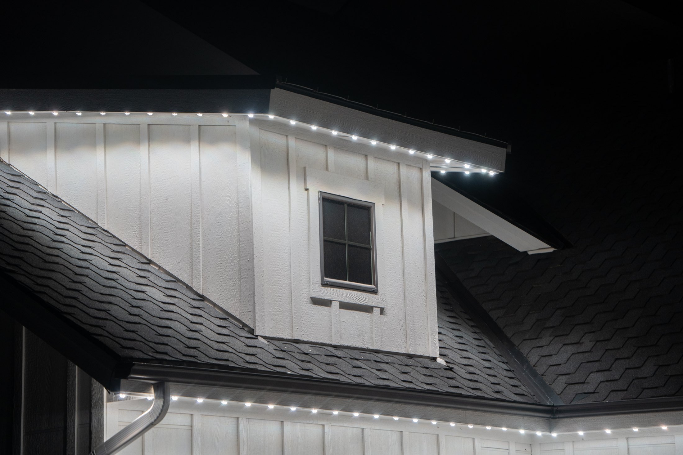White permanent lights follow the upper eaves and lower roof edge of a home, illuminating its white siding at night.
