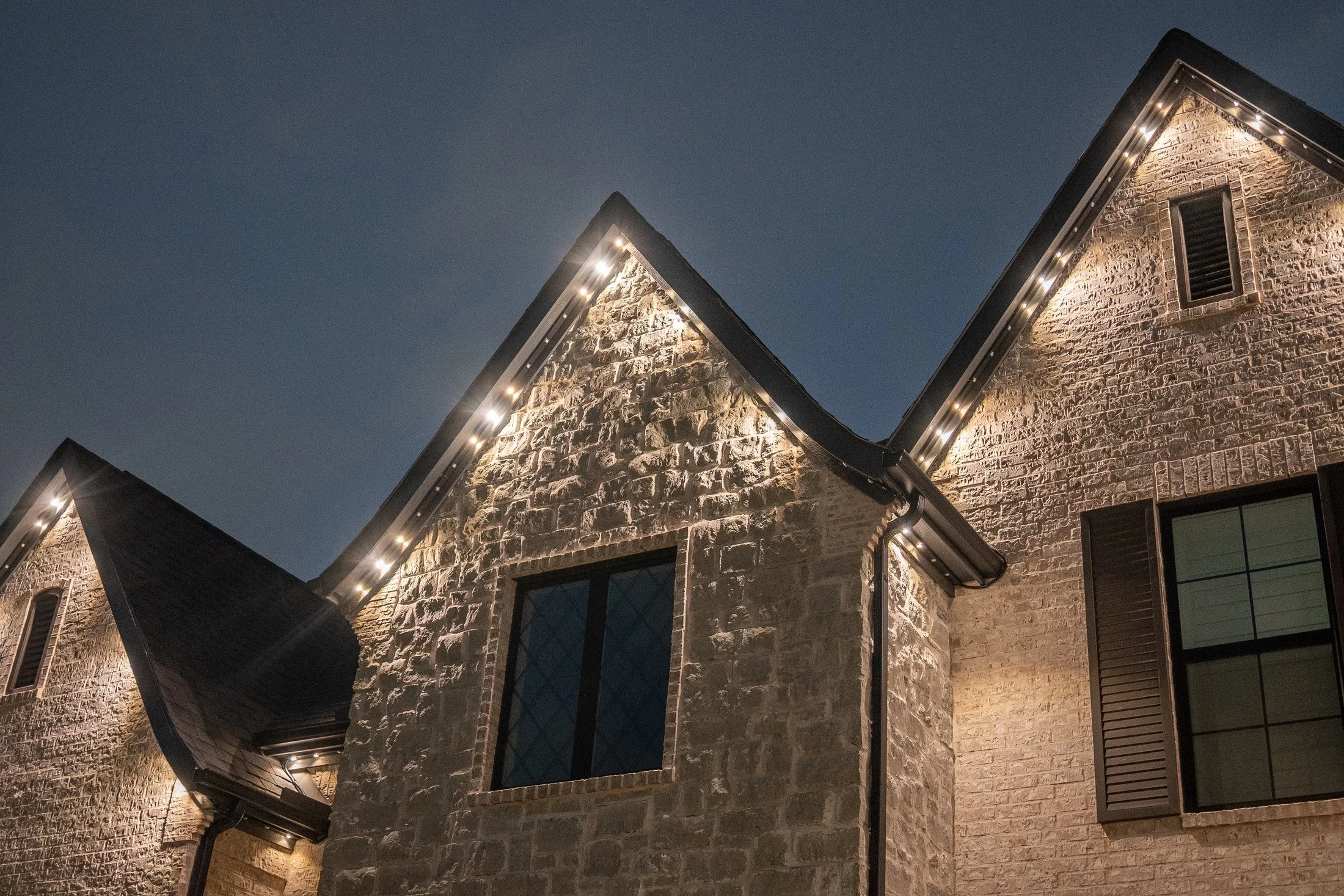Close-up of white lights beneath a stone home’s gable, highlighting the textured masonry.