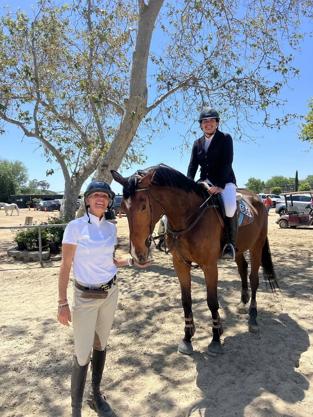A woman and a young rider on a horse at an outdoor equestrian event on a sunny day.