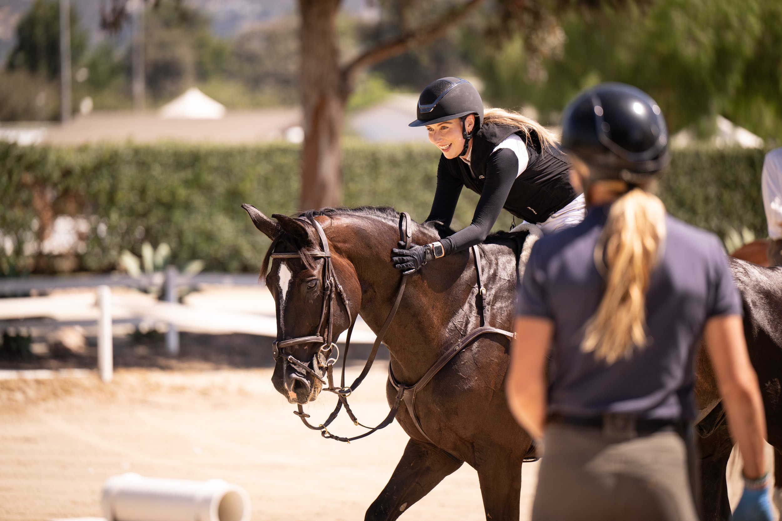 A female rider wearing a black helmet and riding gear, smiling while riding a brown horse in an outdoor equestrian facility. another person in a black helmet and a person in a white shirt are nearby.