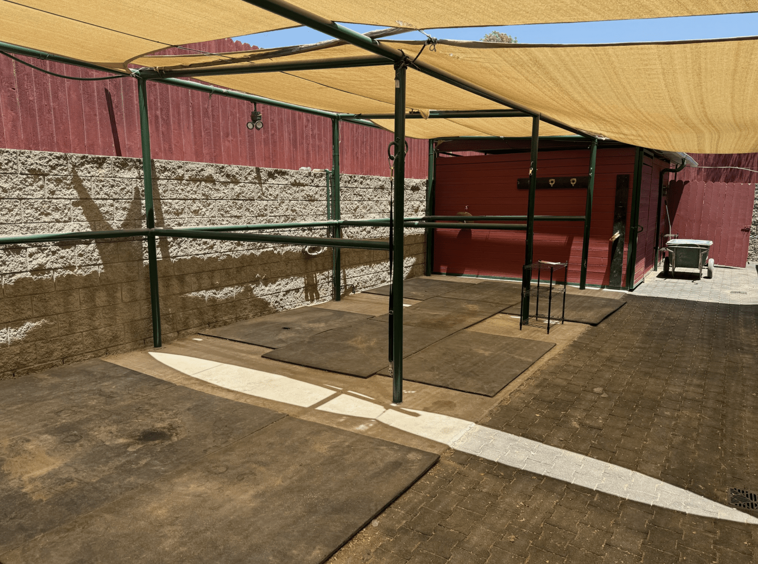 An outdoor dog wash station with a shaded canopy, rubber mats, and a shed with a sink and hoses. There is a wheelbarrow nearby and a red fence in the background.