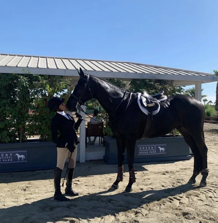 Person in riding attire standing next to a black horse at the desert international horse park.