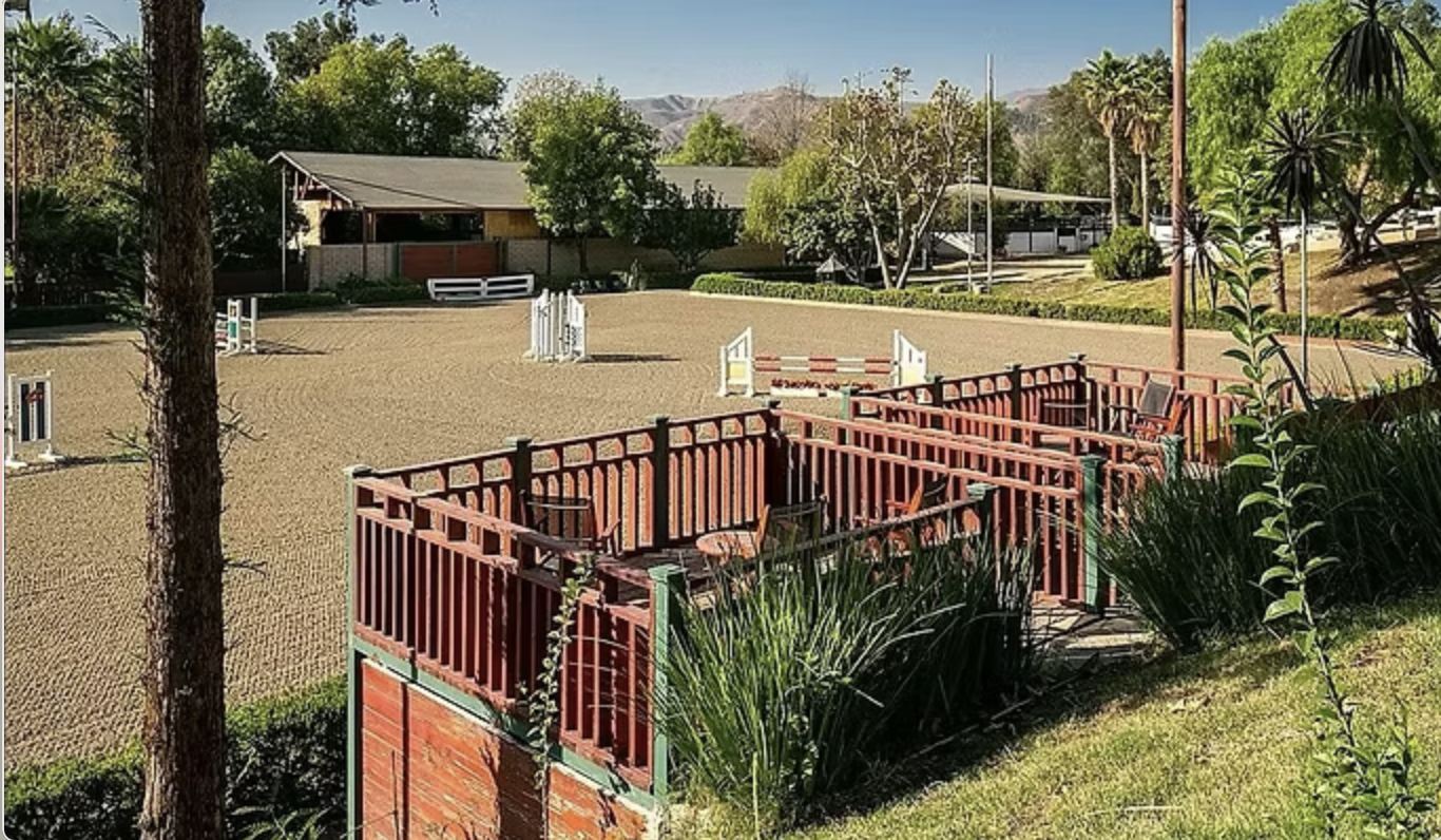An outdoor horse jumping arena with various colorful jumps, surrounded by trees, bushes, and a wooden viewing deck with chairs.