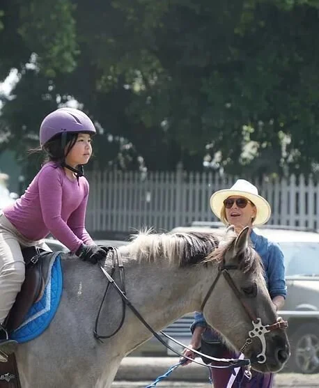 A young girl wearing a purple long sleeve shirt, helmet, and gloves riding a gray horse, with an older woman in sunglasses and a straw hat standing next to them in an outdoor setting.