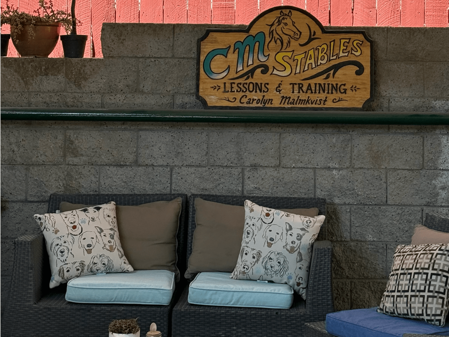 A cozy seating area with a gray wicker sofa adorned with pillows featuring dog illustrations. Behind the sofa, there's a stone wall with a wooden sign that reads 'CM Stables Lessons & Training Carolyn Malmkvist' and has a horse illustration. On top o