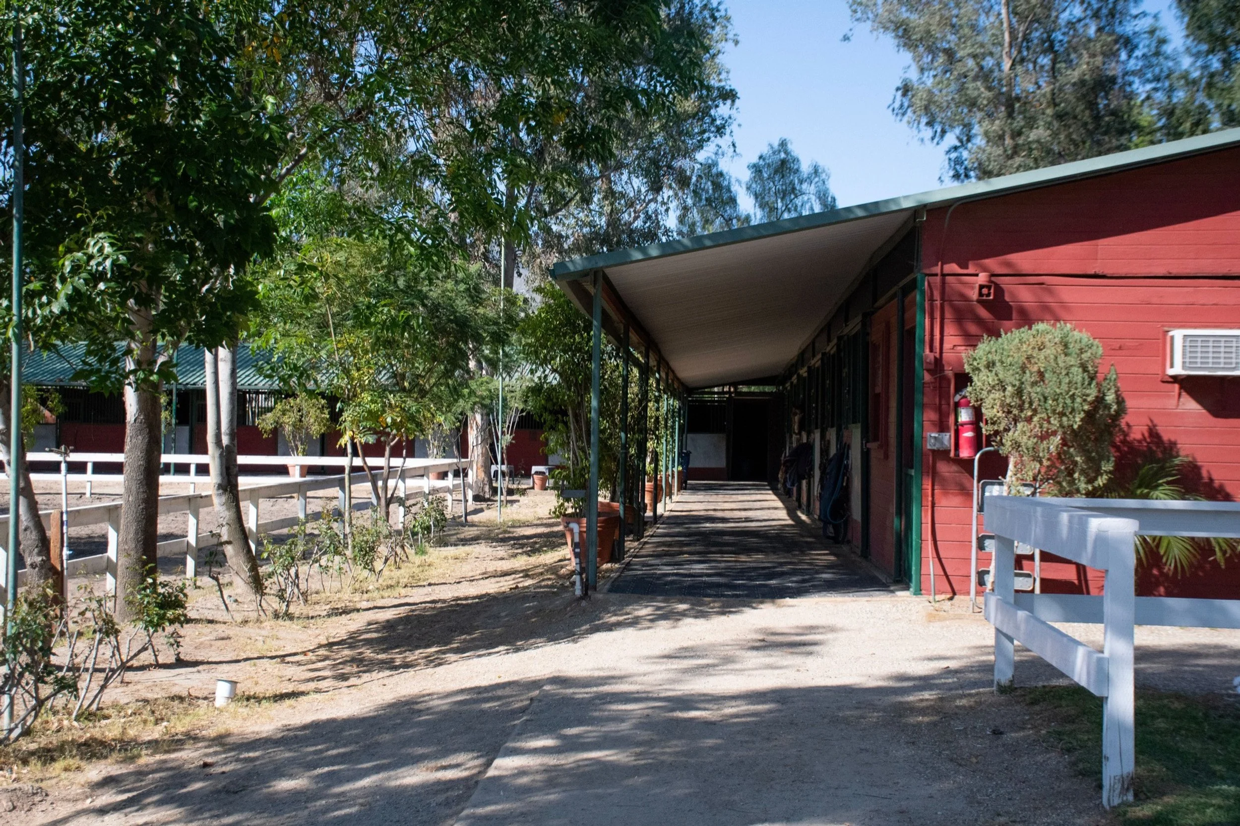 A red building with a covered porch, trees, and a white fence surrounding a dirt area.