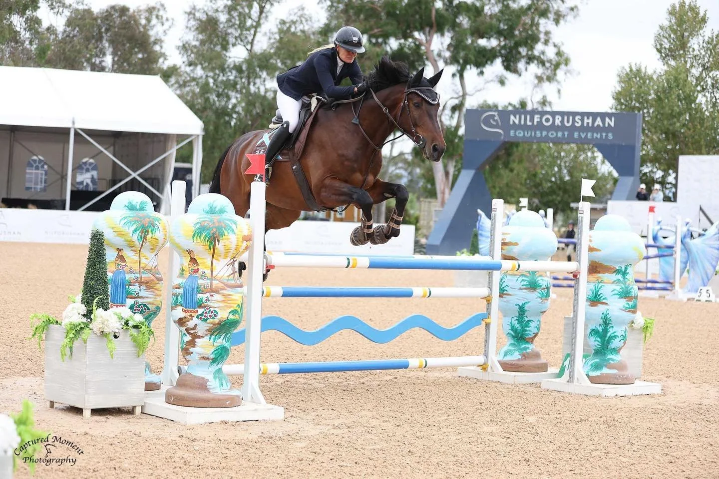 A female equestrian rider on a brown horse jumping over colorful show jumping obstacles with painted vases and greenery at an outdoor competition.