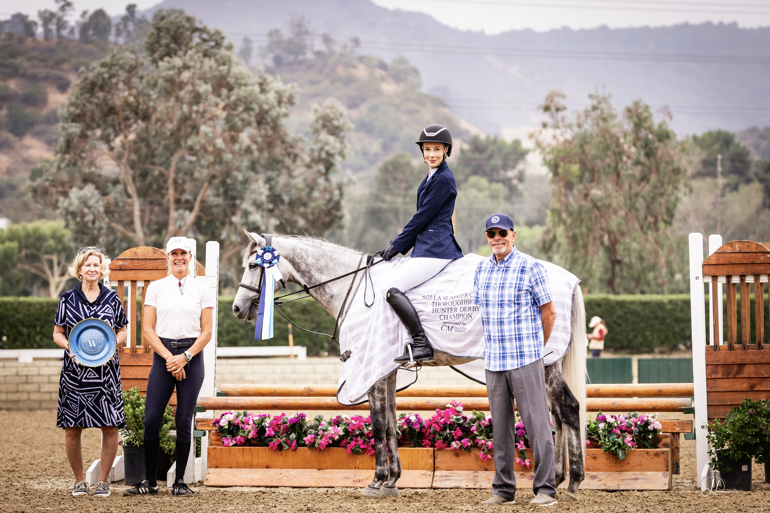 A young woman in a riding helmet and blue blazer riding a gray horse at a horse jumping event, surrounded by three adults, with a scenic background of trees, hills, and mountains.