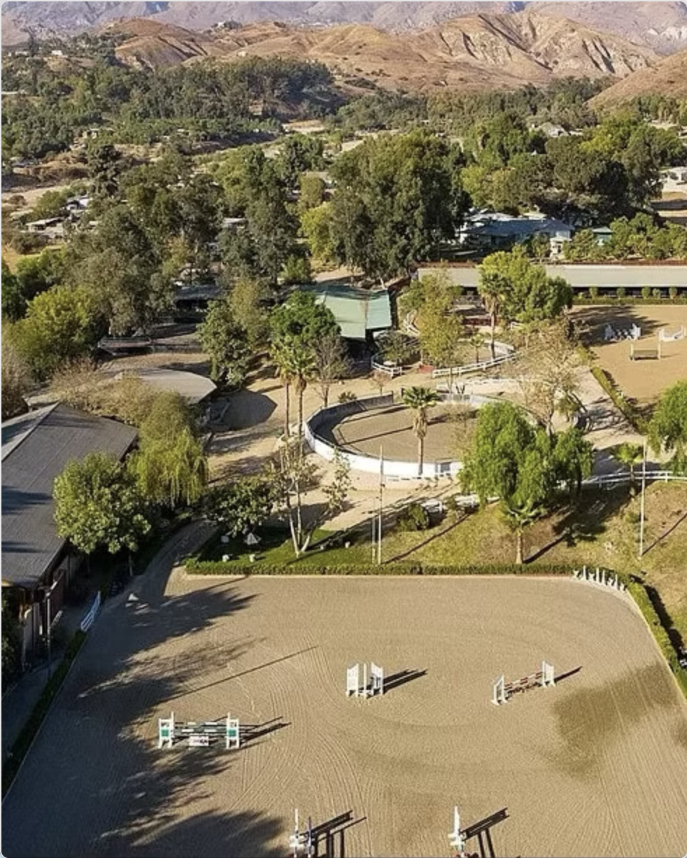 An aerial view of an outdoor equestrian jumping arena with several jumps, surrounded by trees and buildings, with hilly mountains in the background.