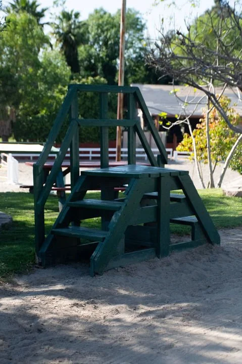 Green wooden playground stairs with handrails on a sandy ground in a backyard with trees and a house in the background.