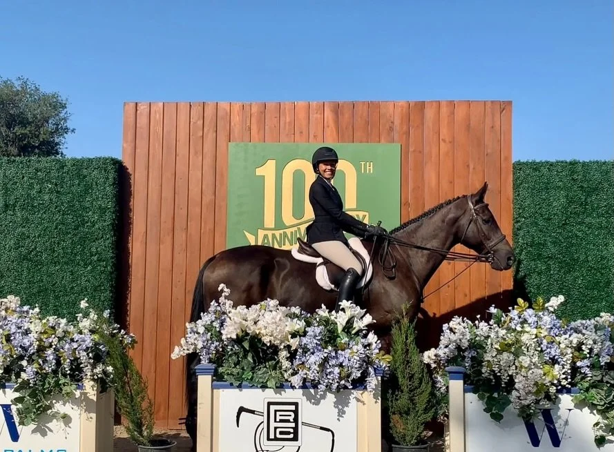 A woman dressed in riding equestrian attire, including helmet and beige riding pants, is sitting on a brown horse decorated with flowers, in front of a wooden fence and a sign celebrating a 100th anniversary.