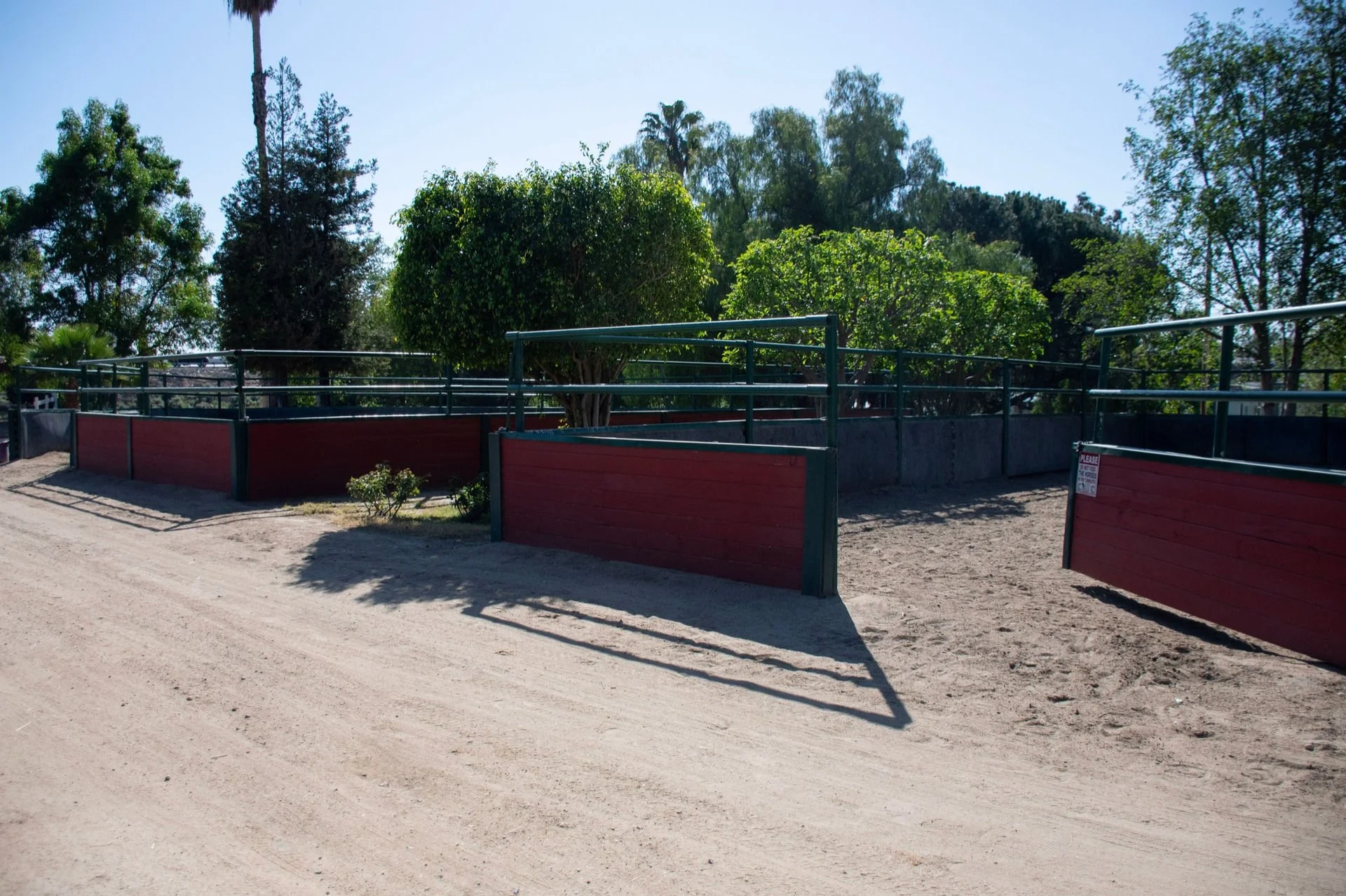 Entrance gate of a horse riding or training area, with red barriers and metal railings, surrounded by trees under a clear sky.