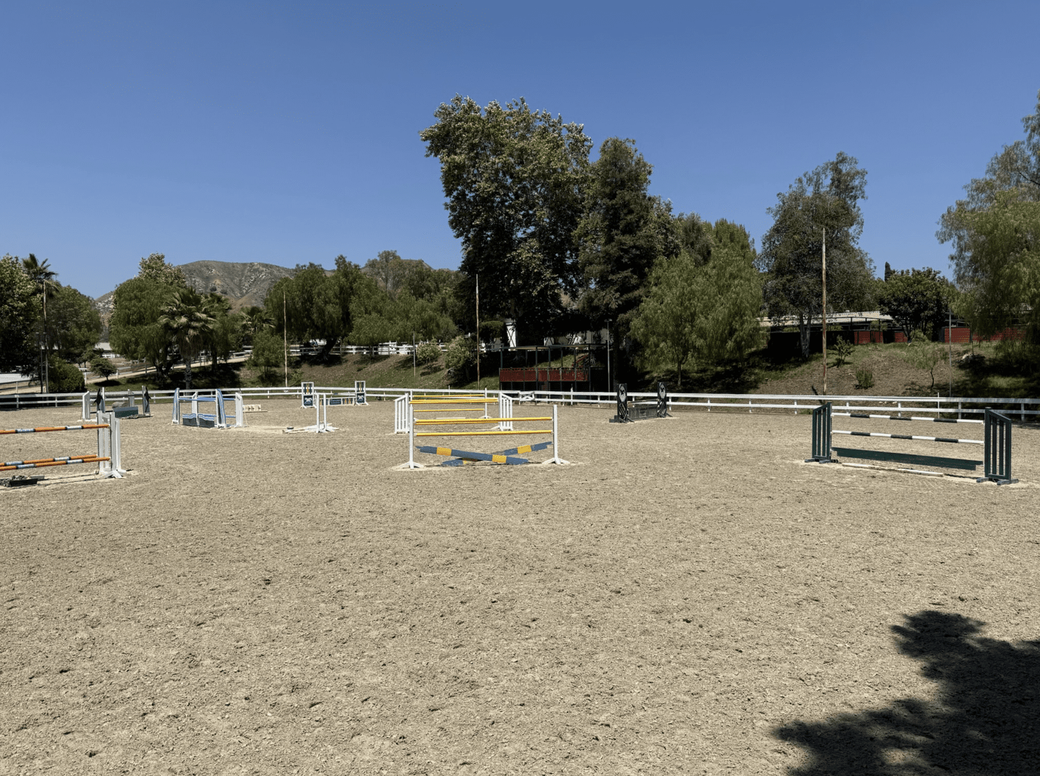 An outdoor horse jumping arena with various colored jumps on a sunny day, surrounded by trees and hills.
