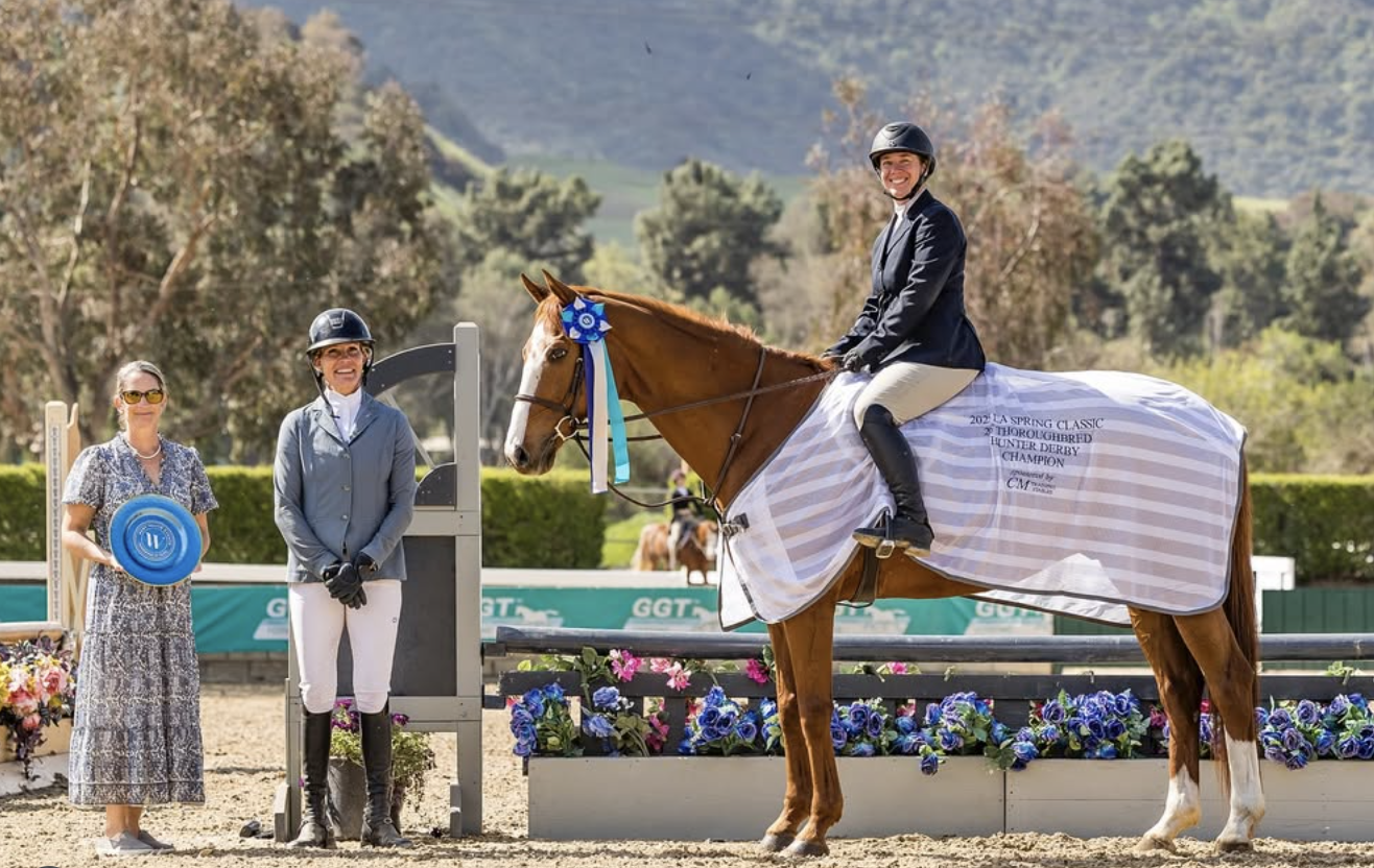 A woman on a brown horse with a blue ribbon, a rider in equestrian riding attire, and two women standing beside them in a competition setting with colorful flowers and hills in the background.