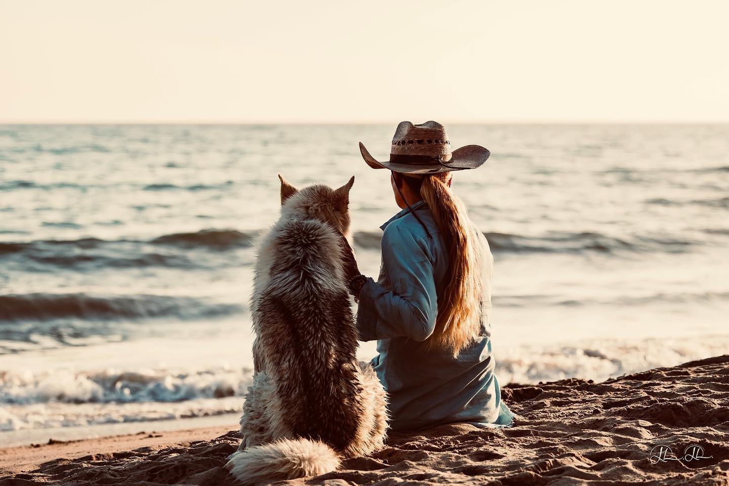A woman wearing a cowboy hat and blue jacket sits on the beach with her dog, a Siberian Husky, as they watch the ocean at sunset.