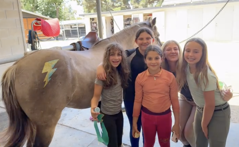 Five smiling girls and a horse inside a stable, with a red saddle and a yellow lightning bolt painted on the horse's side.