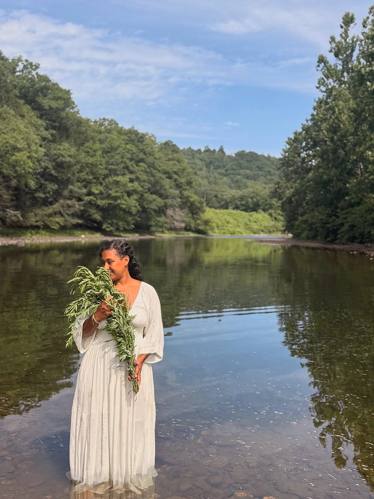 A woman in a white flowing dress standing in a river, holding a large plant with green leaves, with a background of trees and a cloudy sky.