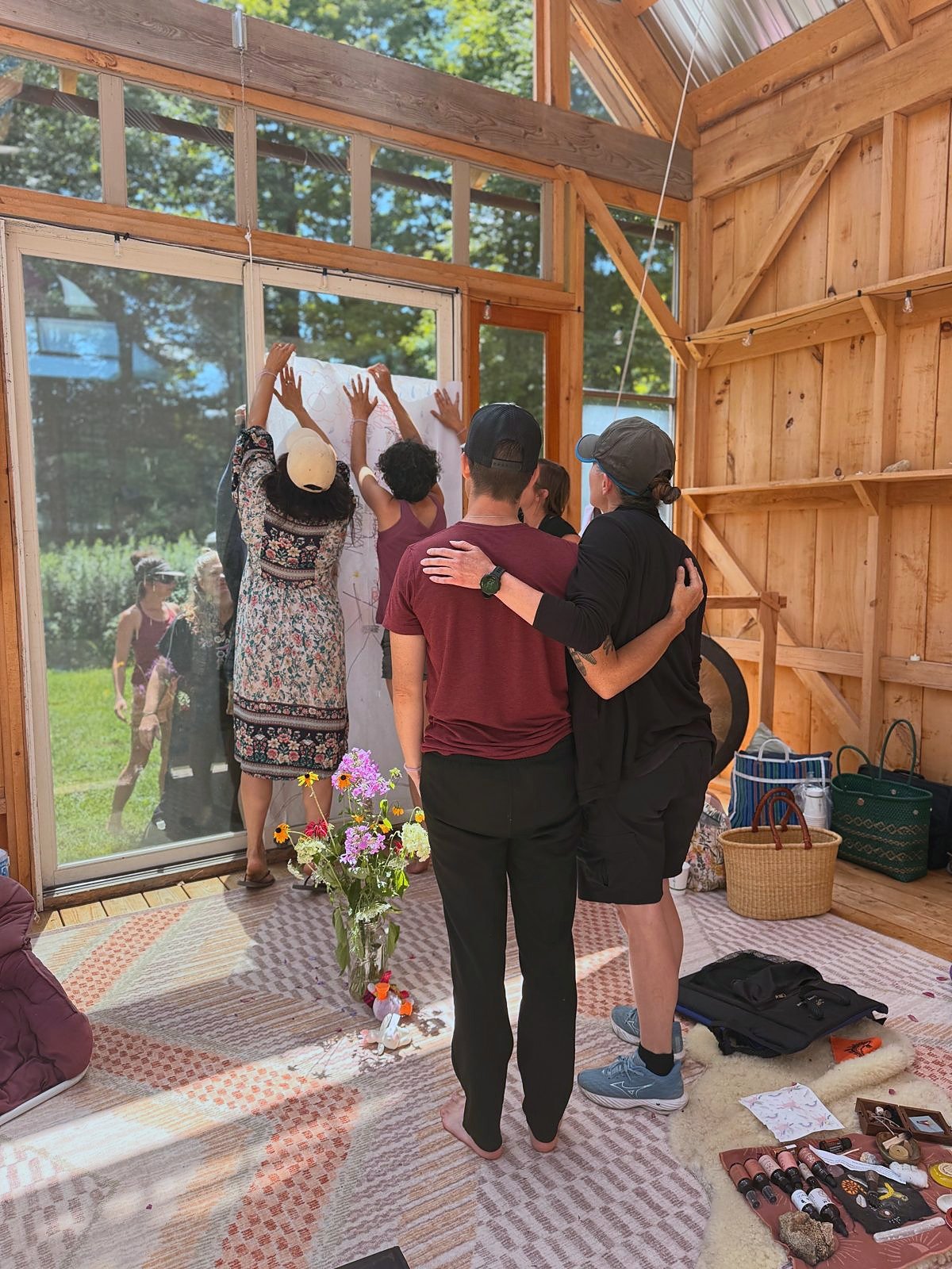 People gathered inside a wooden cabin with large windows, some reaching up to a glass wall, and others embracing, participating in a group activity. There are flowers, makeup, bags, and other personal items on the floor.