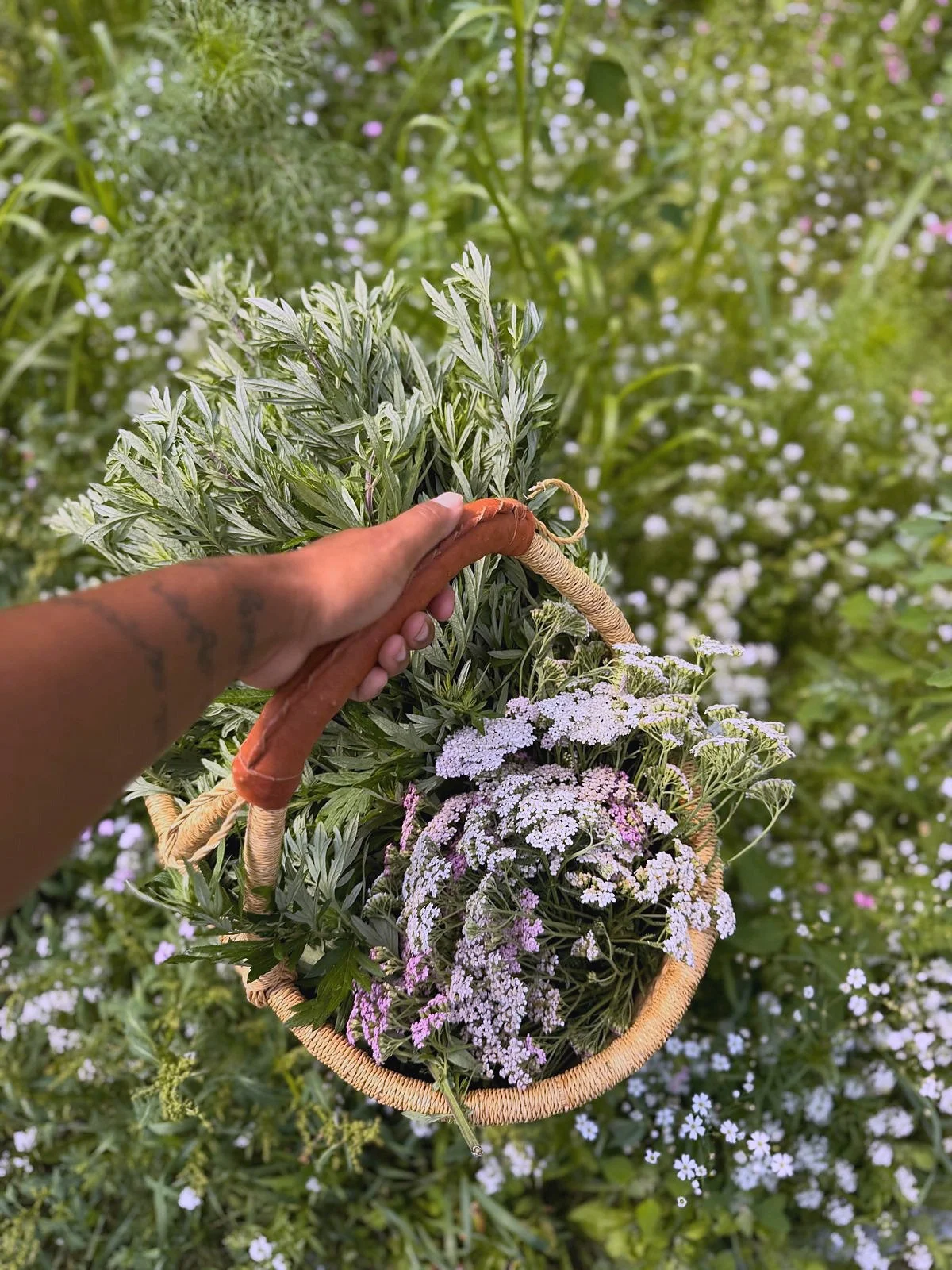 Amber Arnold holding a wicker basket filled with white, pink, and lavender flowers outdoors, with lush green plants and small white flowers in the background.