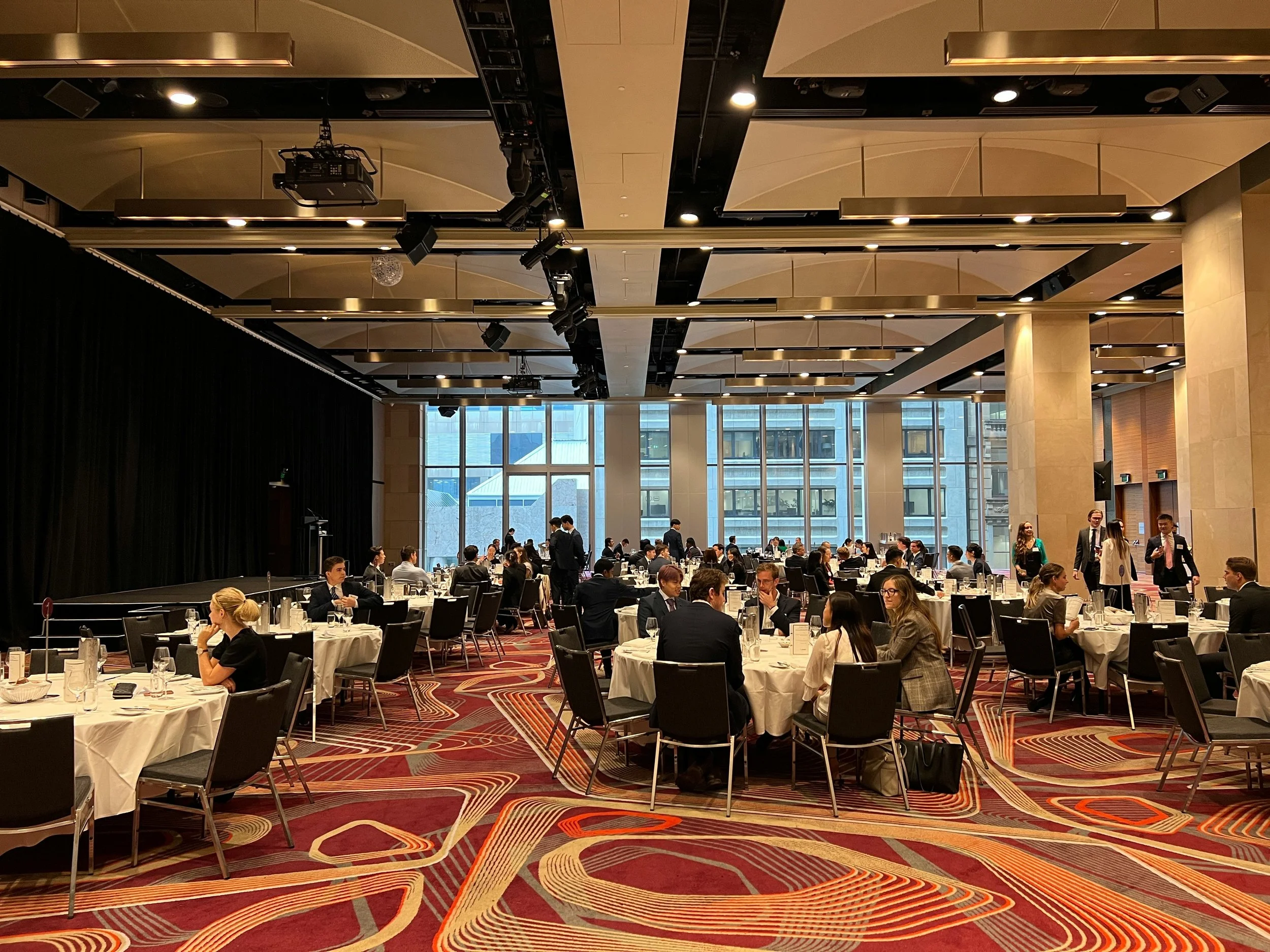 Formal banquet in a hotel ballroom with guests seated at white-clothed tables, a stage, and ceiling-mounted event lighting
