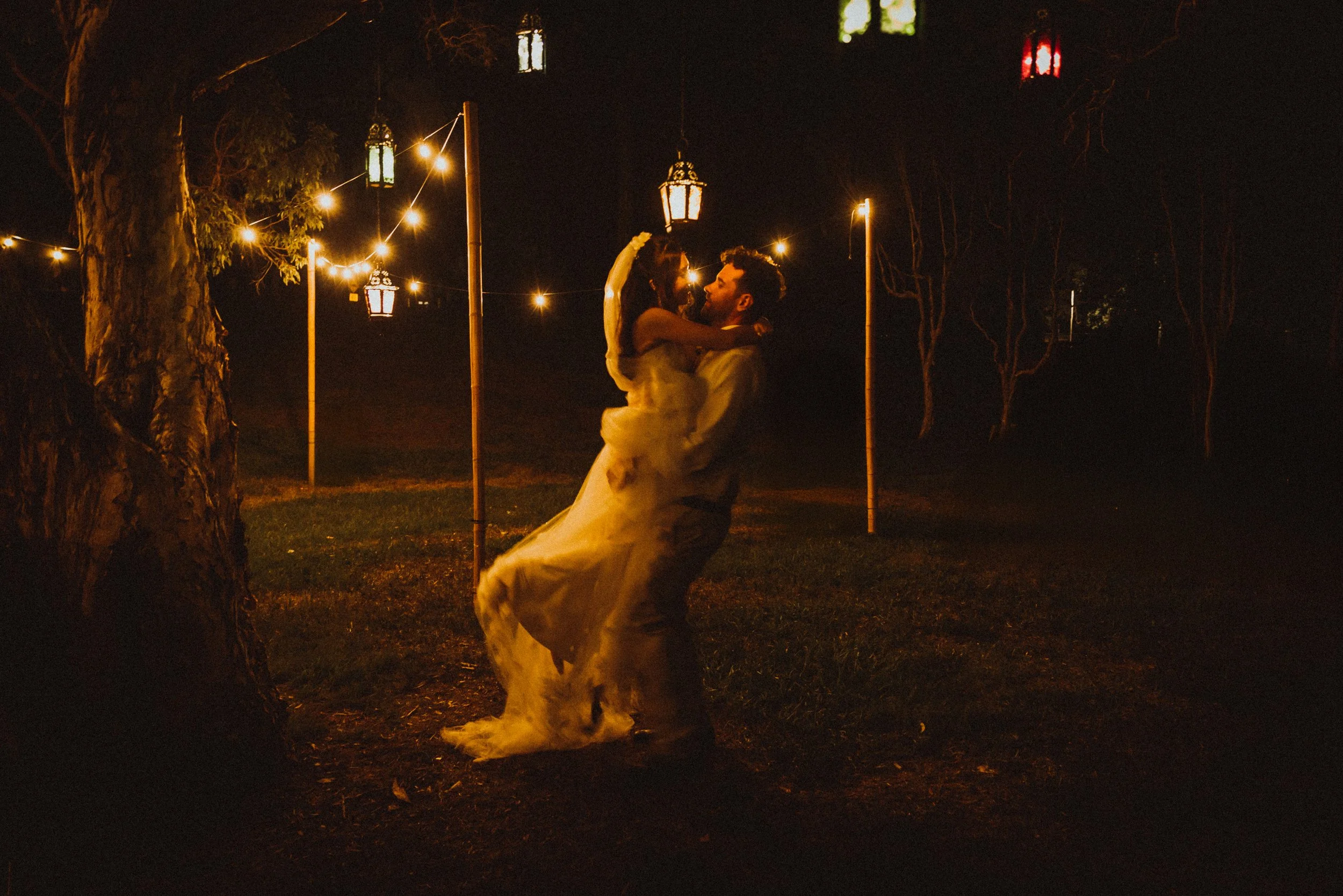 A couple dancing outdoors at night under hanging lanterns and string lights, with trees and lit lampposts in the background.
