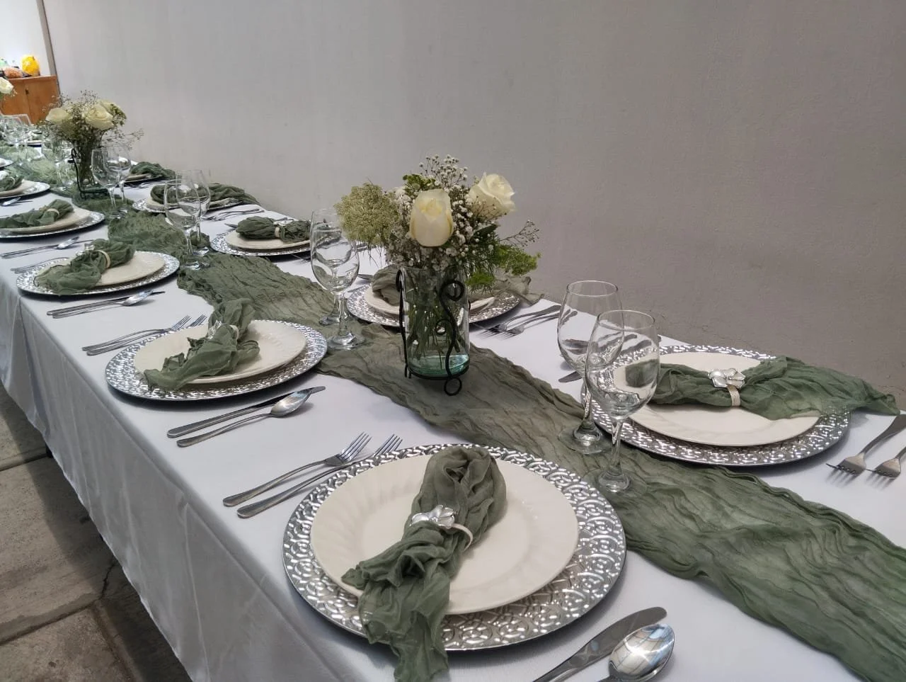 Mesa decorada con mantelería blanca, caminos de tela verde, platos plateados, copas de agua y vino, cubiertos de plata, servilletas verdes enrolladas con anillos blancos, centro de mesa con flor de rosas blancas y pequeñas flores, en un ambiente elegante.
