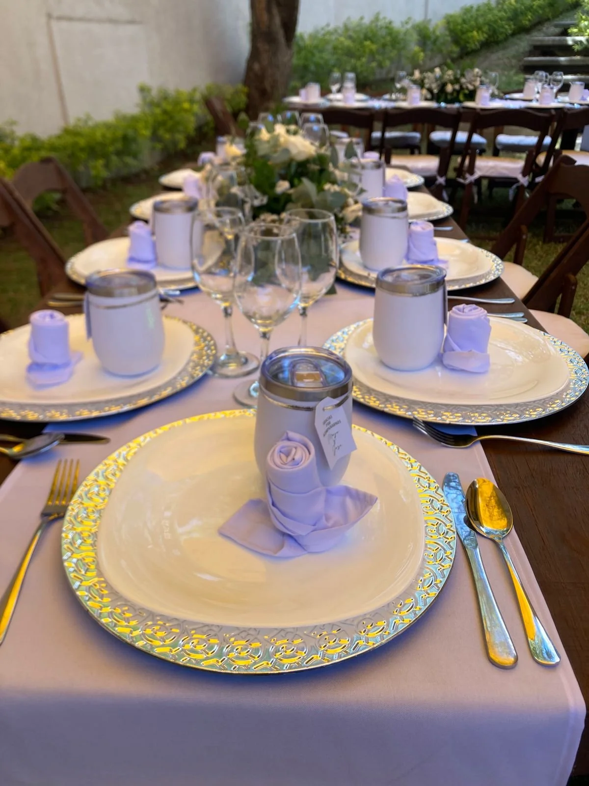 Mesa decorada para evento con platos, copas, cubiertos y vasos, centro de mesa con flores blancas y velas, en un ambiente al aire libre