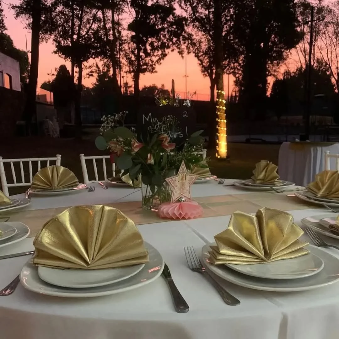 Mesa decorada para una celebración al atardecer con mantelería blanca, platos blancos, cubiertos y servilletas doradas dobladas. En el centro hay un arreglo floral con rosas y follaje, y adornos decorativos en forma de estrella en tonos rosados y dorados. En el fondo, árboles y una puesta de sol rosa y naranja.