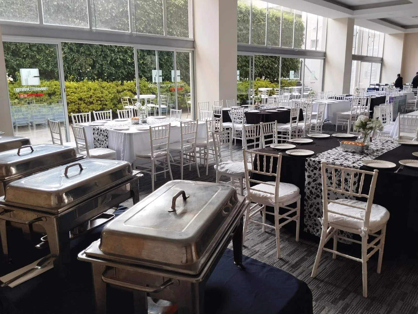 Sala de eventos con mesas y sillas blancas y negras, prepara para una fiesta o reunión, con ventanas grandes que dejan entrar luz natural, y utensilios de comida en la esquina izquierda.
